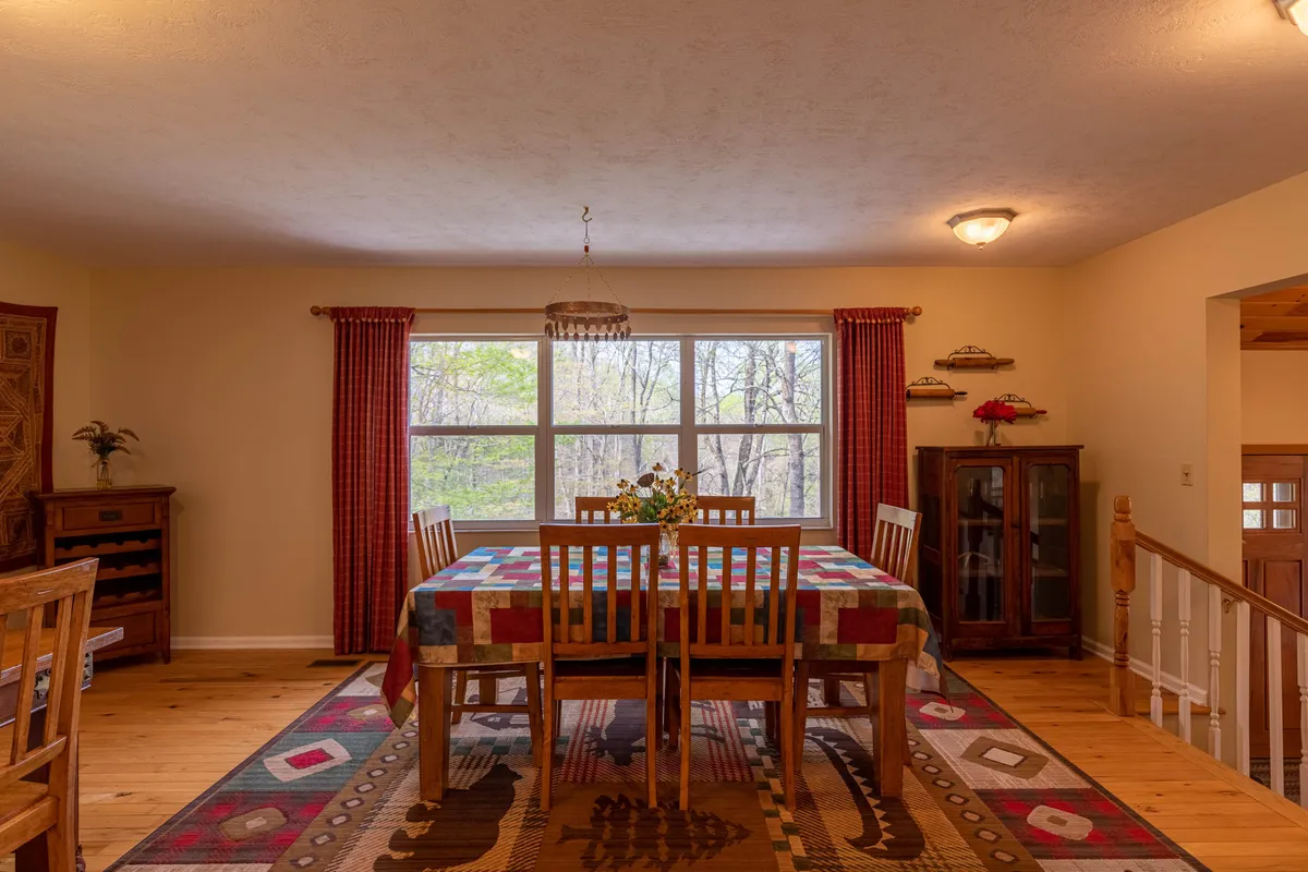 A dining table is set beside expansive windows draped with decorative curtains, offering tranquil forest views. Wooden furniture and warm tones create an inviting ambiance, perfect for family gatherings or quiet breakfasts surrounded by nature.