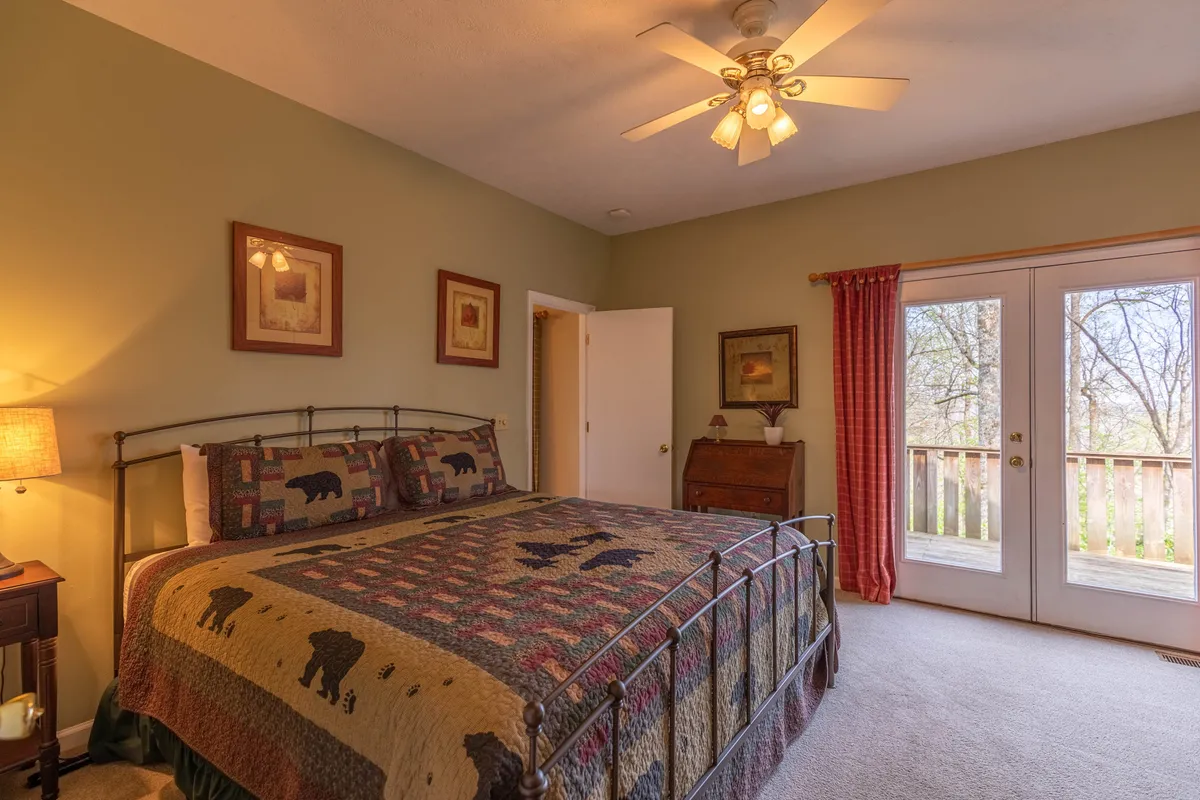 This image features a bed with a patterned quilt, double doors opening to a balcony, and a wooden dresser. The scene is filled with warm, natural light, creating a restful and inviting atmosphere ideal for unwinding. The presence of the balcony hints at private deck access, aligning with the master suite description.
