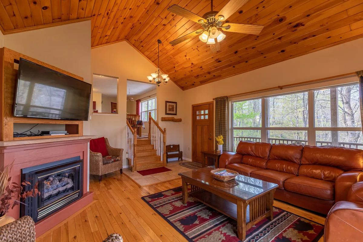 A warm and inviting living area featuring a plush leather sofa, a glowing gas fireplace, expansive windows that frame treetop views, and a rustic wooden ceiling. The space is ideal for relaxing after outdoor adventures or gathering with loved ones during chilly evenings.
