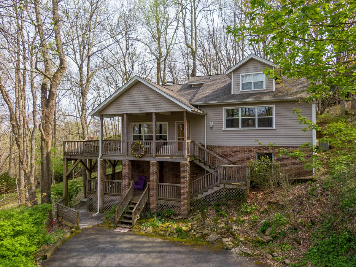 The image shows a charming single-family house nestled among towering trees, with a spacious front porch accessed by wooden steps and large windows that reflect the surrounding forest. The scene conveys a peaceful, secluded atmosphere perfect for relaxing outdoors, with the porch inviting guests to enjoy sunset views or morning coffee while immersed in nature.