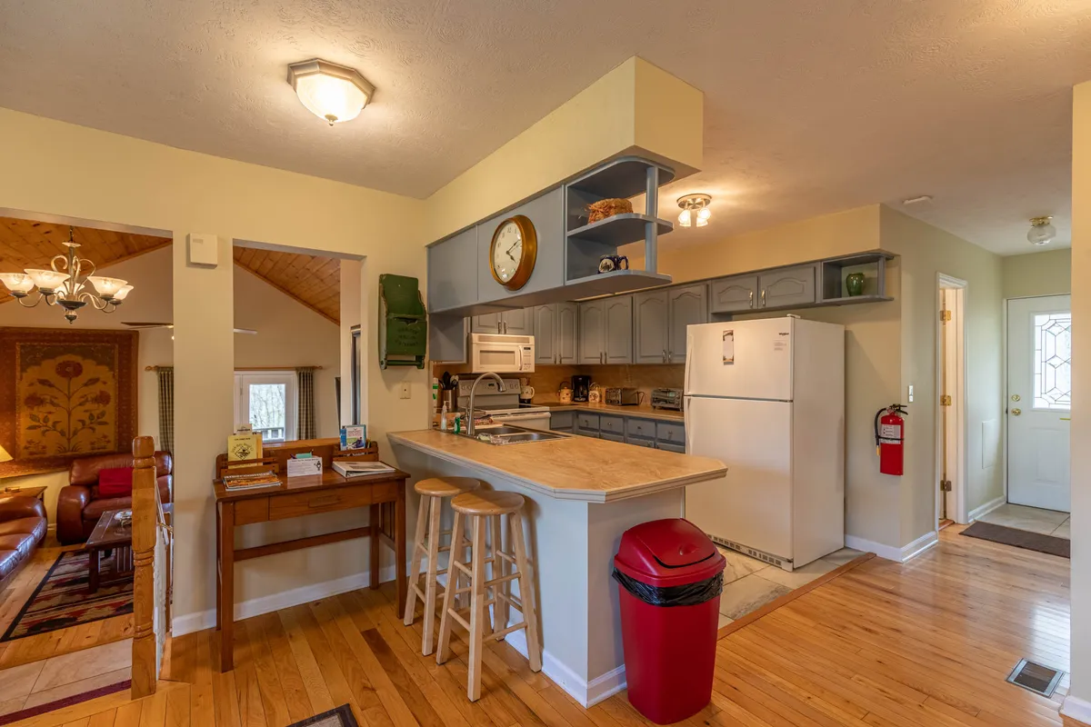 A spacious kitchen and living area featuring a central island with wooden bar stools, a clock on the wall, and a glimpse into the comfortable living space beyond. The area is filled with warm light, inviting for cooking, entertaining, or enjoying family meals together.