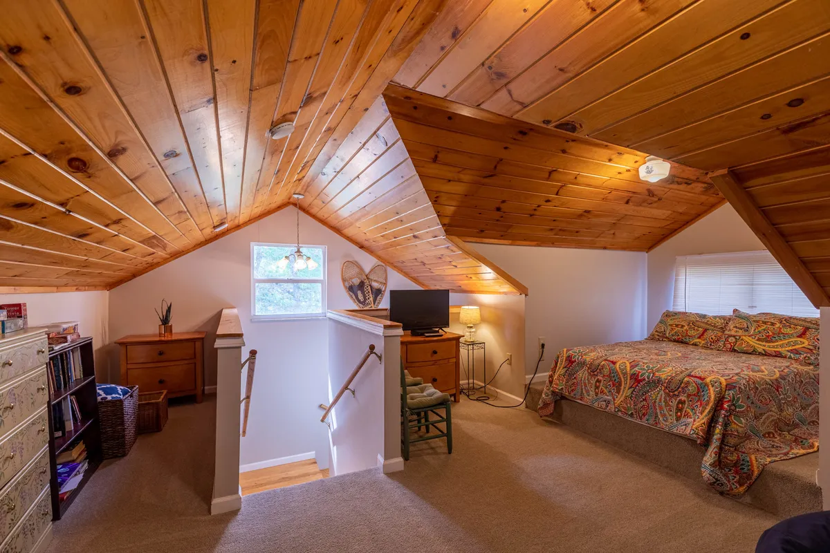 A loft space under a wooden ceiling featuring a colorful bedspread, bookshelf, TV setup, and a staircase. It's both open and cozy, with inviting elements for relaxing, reading, or watching TV—ideal for children, teens, or friends.