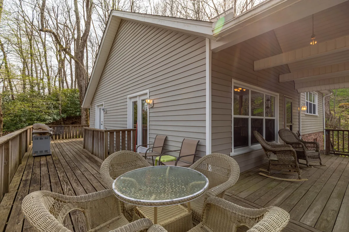 A spacious outdoor deck with a glass-top table surrounded by comfortable chairs and nestled among tall trees. The area is designed for relaxation and dining, with the natural beauty of the forest visible in every direction. The morning light filters softly through the branches, creating a serene and inviting gathering spot for families.