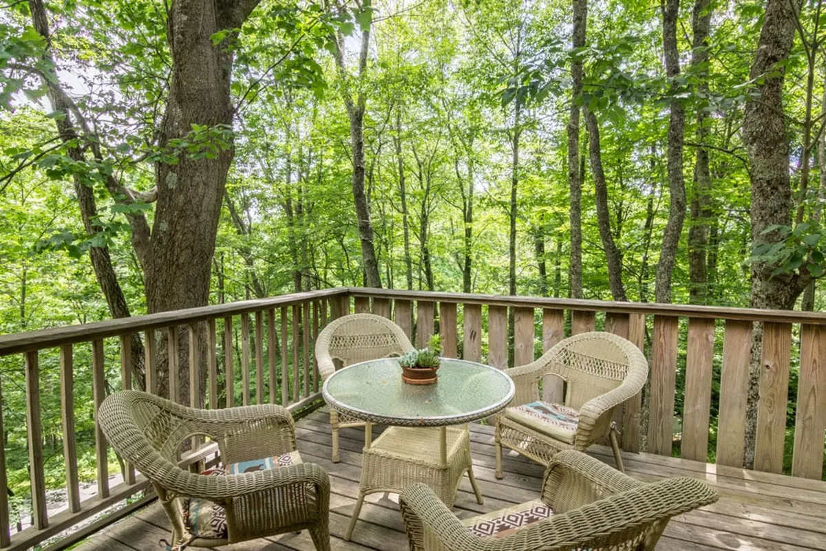 This image features wicker chairs and a round glass table set on an outdoor deck, surrounded by lush green trees. The scene conveys a peaceful ambiance with plenty of natural light, suggesting a perfect spot for sipping coffee or reading while immersed in the sights and sounds of nature.