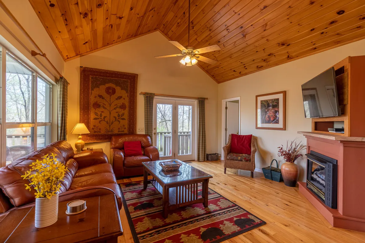 This image showcases the living room area, featuring supple leather couches arranged around a stone fireplace, with a wooden ceiling overhead and decorative wall art. The coffee table and ambient lighting enhance the sense of warmth and comfort, inviting relaxation or socializing in an intimate setting.